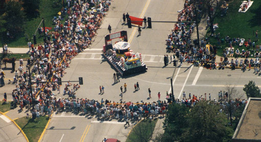 Aerial photograph of a parade float rounding a corner. People line the streets to see the parade.
